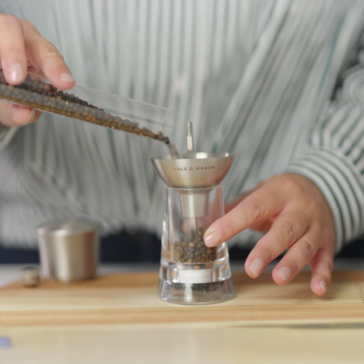 A person pours black peppercorns from a small container into a glass pepper grinder with a metal top, standing on a wooden surface.