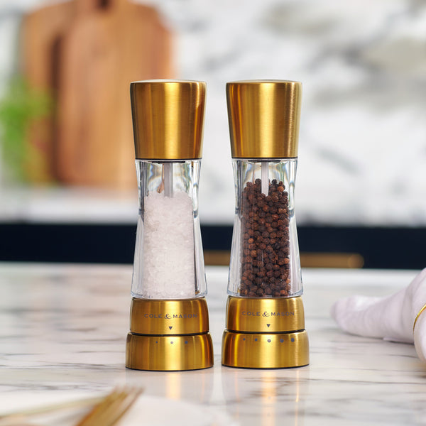 Modern Cole & Mason gold stainless steel and acrylic salt and pepper grinders with adjustable grind settings on a kitchen counter