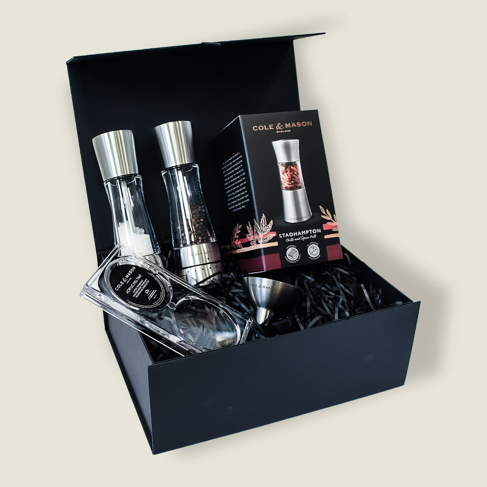 The Derwent Mill Christmas Gift Hamper by Cole & Mason features salt and pepper mills, a metal funnel, and a branded box with spice grinder image—all presented in black shredded paper. Perfect as a festive gift hamper.