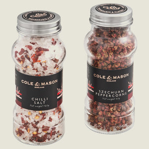 The Cole & Mason Aromatic Salt & Peppercorns Gift Set features two clear jars with silver lids, each labeled in black with red and white accents—one contains chili salt and the other Szechuan peppercorns.