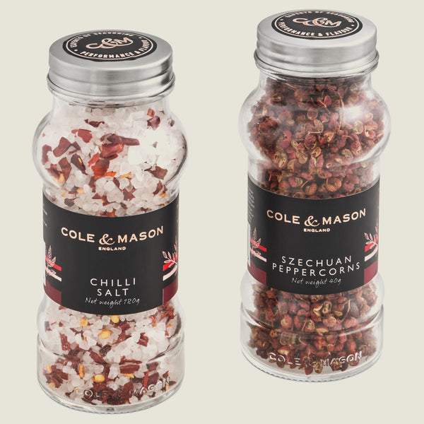 The Cole & Mason Aromatic Salt & Peppercorns Gift Set features two clear jars with silver lids, each labeled in black with red and white accents—one contains chili salt and the other Szechuan peppercorns.