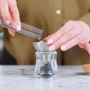 A person pours whole peppercorns into a glass pepper grinder, using a metal funnel, on a white marble countertop. The person is wearing a brown shirt.