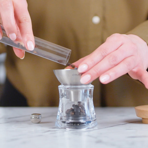 A person pours whole peppercorns into a glass pepper grinder, using a metal funnel, on a white marble countertop. The person is wearing a brown shirt.