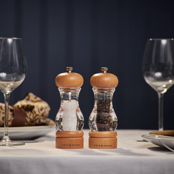 The Cole & Mason 105 Beech Salt & Pepper Mills, with clear bodies and beech wood tops and bases plus adjustable grinding, are displayed on a set dining table with empty wine glasses and plates in the background.