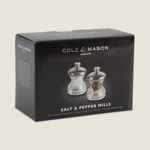 A black box displays the Cole & Mason Button Salt & Pepper Mill Set with metallic tops and a compact design. The clear mills are shown, alongside text reading Salt & Pepper Mills and noting adjustable coarseness in smaller font.