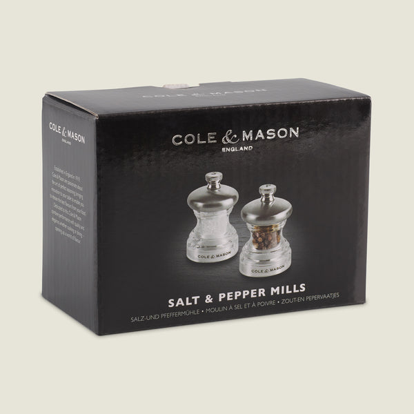 A black box displays the Cole & Mason Button Salt & Pepper Mill Set with metallic tops and a compact design. The clear mills are shown, alongside text reading Salt & Pepper Mills and noting adjustable coarseness in smaller font.