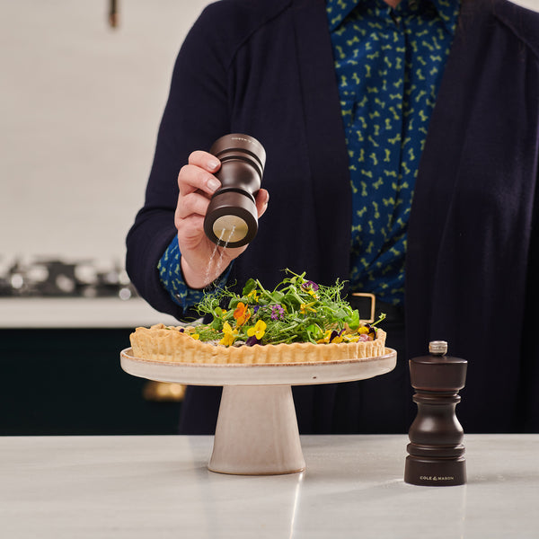 A person sprinkles pepper from the Cole & Mason London Chocolate Wood Pepper Mill onto a vegetable tart with greens and flowers, displayed on a cake stand. The matching Salt Shaker from the set rests on the countertop nearby.