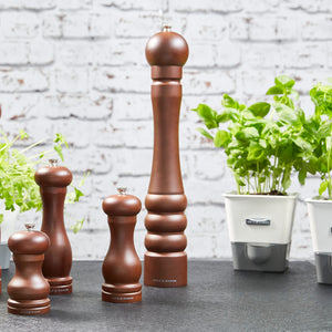 The Cole & Mason Beech Capstan Pepper Mill 400mm, featuring adjustable grind, is displayed on a dark countertop beside potted basil and herbs in white containers, all set before a white brick wall.