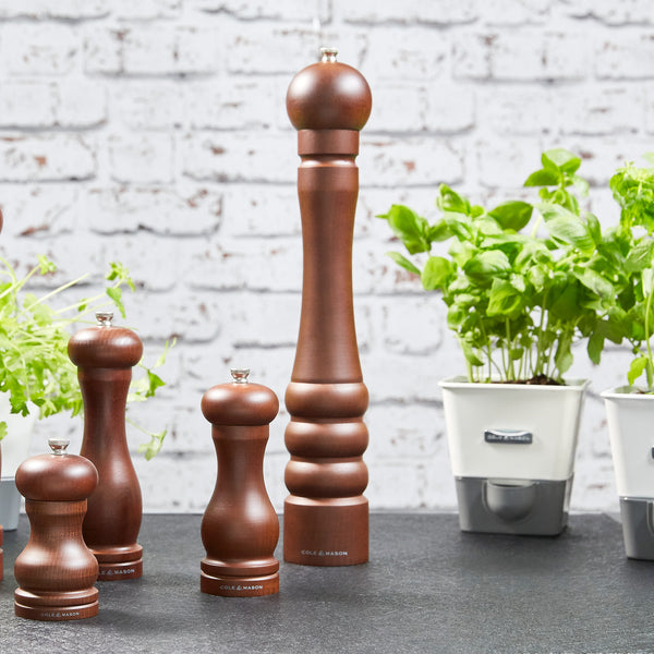 The Cole & Mason Beech Capstan Pepper Mill 400mm, featuring adjustable grind, is displayed on a dark countertop beside potted basil and herbs in white containers, all set before a white brick wall.
