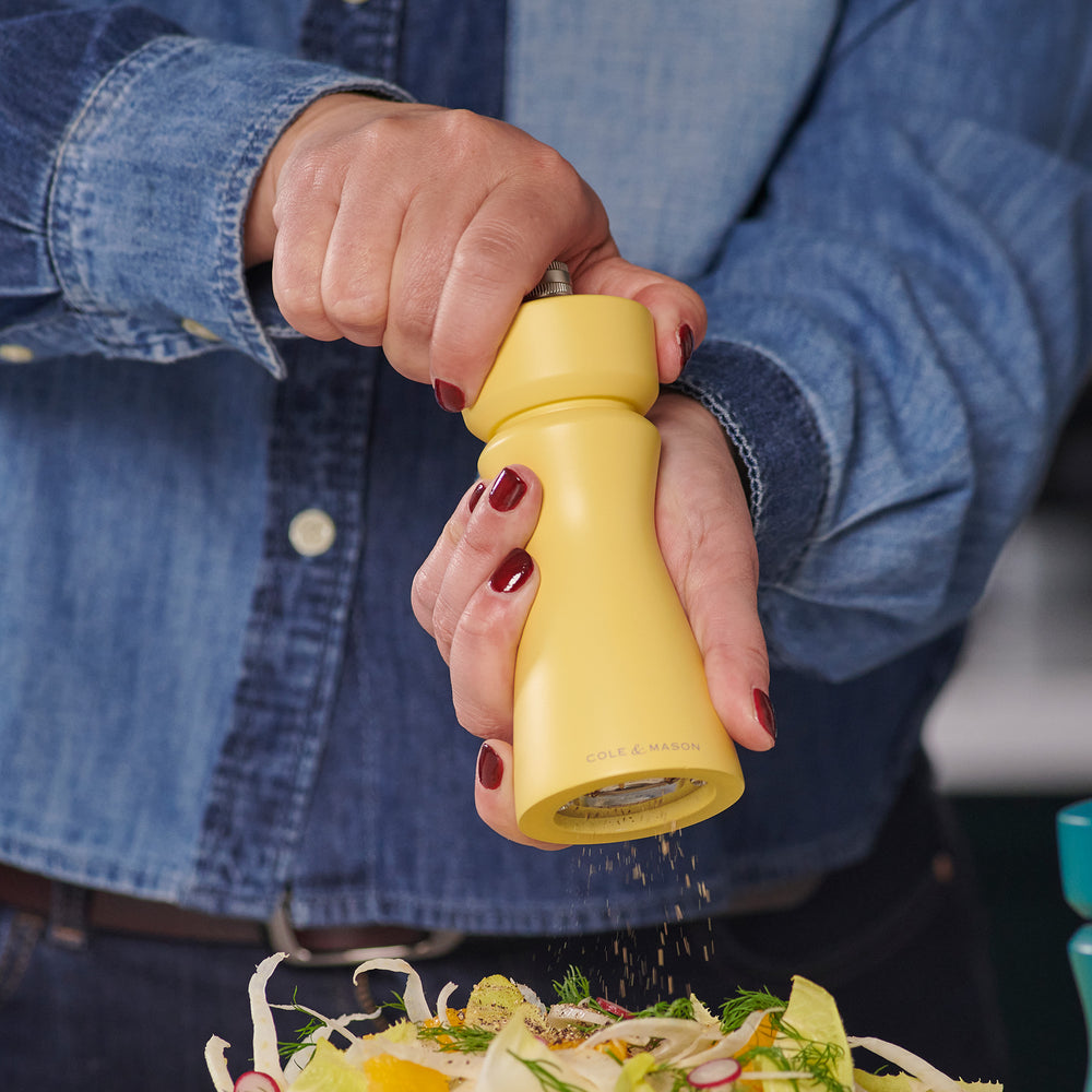 The Cole & Mason Kenton Salt & Pepper Mill in Turmeric Yellow features a silver top knob, brand logo at the base, and a durable ceramic grinding mechanism. Its photographed on a plain light background with a soft shadow to the right.