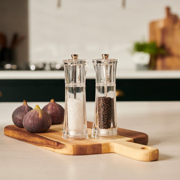 The Cole & Mason UK Prism Salt & Pepper Mill Set Acrylic 165mm, featuring adjustable grind and a stainless steel mechanism, sits on a wooden board next to three whole figs on a kitchen counter with a blurred kitchen background.