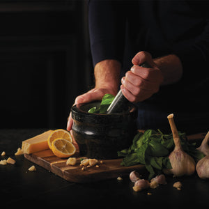 #140mm
A person uses a mortar and pestle to grind fresh basil. Nearby, garlic bulbs, lemon slices, Parmesan cheese, and chopped garlic rest on a wooden cutting board. The scene is dimly lit, emphasizing the ingredients.