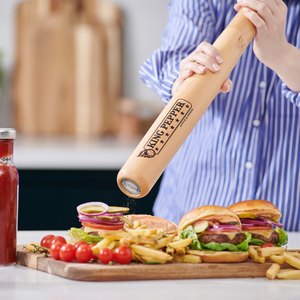 A person seasons burgers and fries with the Cole & Mason King Pepper Baseball Bat Pepper Mill 710mm, which features a carbon steel mechanism and adjustable grind, next to a bottle of ketchup, cherry tomatoes, and food on a white kitchen counter.