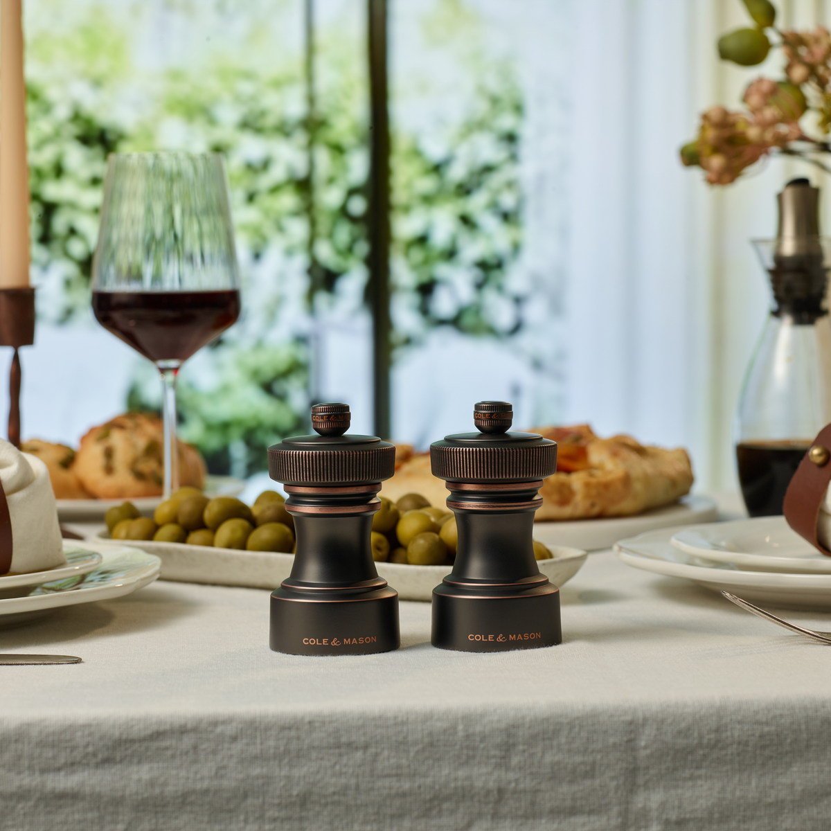 On a set dining table with wine, bread, olives, and plates, the Cole & Mason Hoxton Salt and Pepper Mill Set in Antique Bronze features elegant dark wood and a premium grinding system, positioned in front of a window revealing a lush green outdoor view.