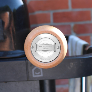 Close-up of the Cole & Mason King Pepper Baseball Bat Pepper Mill 710mm, showcasing its carbon steel mechanism and wooden handle. The blurred background highlights a grill and brick wall, while the mill’s adjustable grind guarantees precise seasoning every time—making it the ultimate King Pepper Mill for your kitchen.