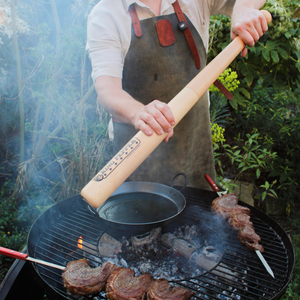 Clad in an apron, a person uses a large wooden rolling pin to press down on a grill pan over the barbecue, where skewered steaks cook amid lush greenery. Within easy reach sits the Cole & Mason King Pepper Baseball Bat Pepper Mill 710mm, poised to add freshly ground flavor to the sizzling steaks.