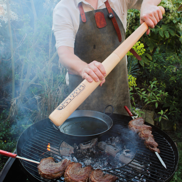 Clad in an apron, a person uses a large wooden rolling pin to press down on a grill pan over the barbecue, where skewered steaks cook amid lush greenery. Within easy reach sits the Cole & Mason King Pepper Baseball Bat Pepper Mill 710mm, poised to add freshly ground flavor to the sizzling steaks.
