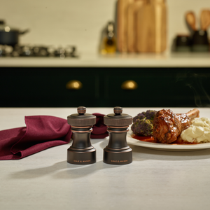 Two dark-colored grinders from the Cole & Mason Hoxton Salt and Pepper Mill Set in Antique Bronze sit on a countertop near a plate of mashed potatoes, broccoli, and meat. A burgundy napkin and kitchen backdrop enhance the scene, highlighting the set’s elegant grinding system—perfect for any dining setting.