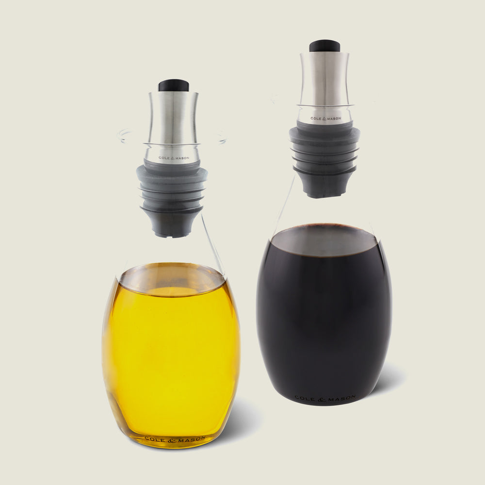 The Cole & Mason UK Haverhill Oil & Vinegar Pourers Set 450ml includes two glass pourers with stainless steel spouts, adjustable rubber stoppers, and a drip return system—one holds yellow oil, the other dark brown vinegar.