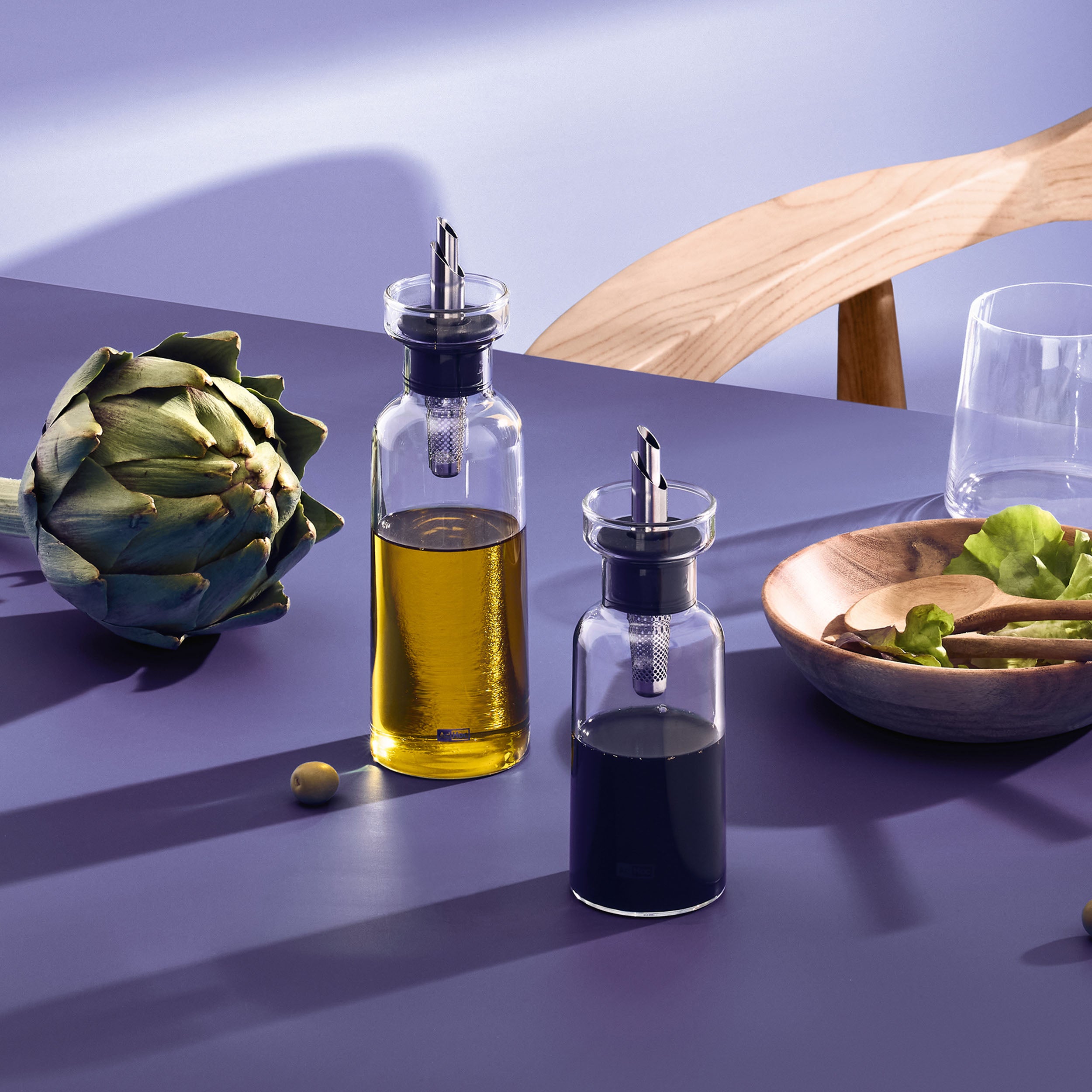 Two glass bottles with metal pour spouts filled with oil and vinegar sit on a purple table near a wooden chair, an artichoke, a glass of water, and a wooden bowl with salad greens.