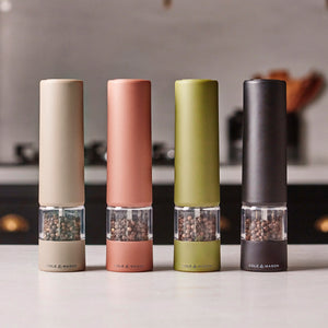 Four sleek Brixton Electric Mills by Cole & Mason, including the Matt Black GS model alongside versions in beige, terracotta, and olive green, are displayed on a kitchen counter—each filled with peppercorns and featuring a durable ceramic grinder. The softly blurred kitchen background enhances their contemporary appeal.