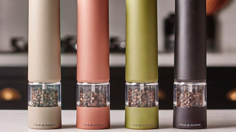 Four sleek, modern Cole & Mason Brixton Electric Mill Stone GS grinders with an adjustable grind and durable ceramic grinding core in beige, peach, olive green, and black stand in a row on a kitchen counter, with a blurred stove and utensils in the background.