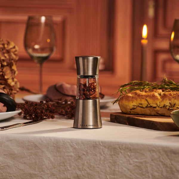 A Cole & Mason Derwent Mill Christmas Gift Hamper sits on a white tablecloth with bread, dried flowers, wine glasses, and a candle—an elegant addition to any festive dining setting.