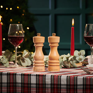 A festive table features a plaid cloth, two red wines, red candles, eucalyptus branches, and the Cole & Mason London Natural Beech Mill Christmas Gift Hamper. Warm lights and greenery glow in the background—perfect for holiday gifting.