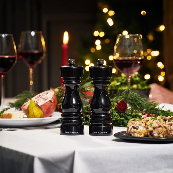 The Cole & Mason London Black Gloss Mill Christmas Gift Hamper sits on a festive table with wine glasses, food, a lit candle, holiday lights, greenery, and flowers—perfect for gifting this season.