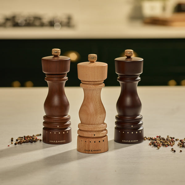 Three Cole & Mason London Walnut Salt or Pepper Mills—two in dark walnut finish and one in a lighter tone—are displayed on a kitchen counter with scattered peppercorns, highlighting the refined design and adjustable functionality of these mills. In the background, a stove and various kitchen items are softly blurred.