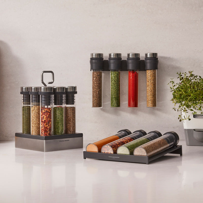 The Cole & Mason Shadwell Slim-Mount Spice Storage features airtight glass containers on a white counter, with tubes displayed vertically and as a wall-mounted rack. A small potted plant sits nearby.