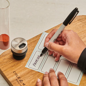 A hand writes PAPRIKA on a label with a black marker. The Shadwell Countertop Spice Storage by Cole & Mason, airtight glass containers, a spice jar lid, and a wooden cutting board sit on a light countertop.