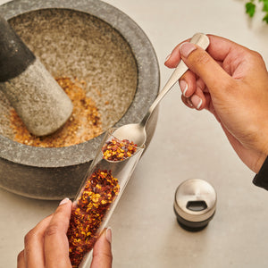 A person scoops dried chili flakes from a Shadwell Countertop Spice Storage by Cole & Mason, beside a stone mortar and pestle—ideal for keeping your spices organized on the countertop.