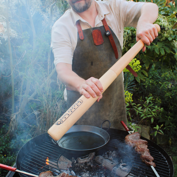 Wearing a brown apron, someone uses the Cole & Mason King Pepper Baseball Bat Pepper Mill 710mm to season food on an outdoor barbecue grill, surrounded by greenery and smoke.