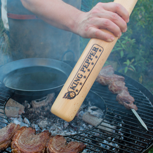 Grilling skewers and steaks over charcoal, a person uses the Cole & Mason King Pepper Baseball Bat Pepper Mill 710mm, with smoke rising and a cast-iron pan in the background—demonstrating its versatile adjustable grind.