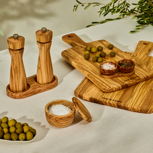 The Cole & Mason Portobello Set Bundle with M Board features wooden salt and pepper mills, an Italian olive wood board, green olives, chopped herbs, and a lidded salt cellar on a cream tablecloth with olive branches in the background.