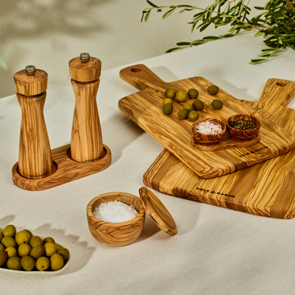 The Cole & Mason Portobello Set Bundle with M Board features wooden salt and pepper mills, an Italian olive wood board, green olives, chopped herbs, and a lidded salt cellar on a cream tablecloth with olive branches in the background.