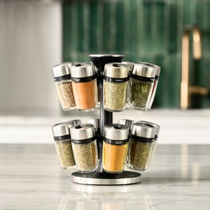 A rotating spice rack with glass jars containing various spices sits on a marble countertop. The background features green tile and a blurred gold faucet.