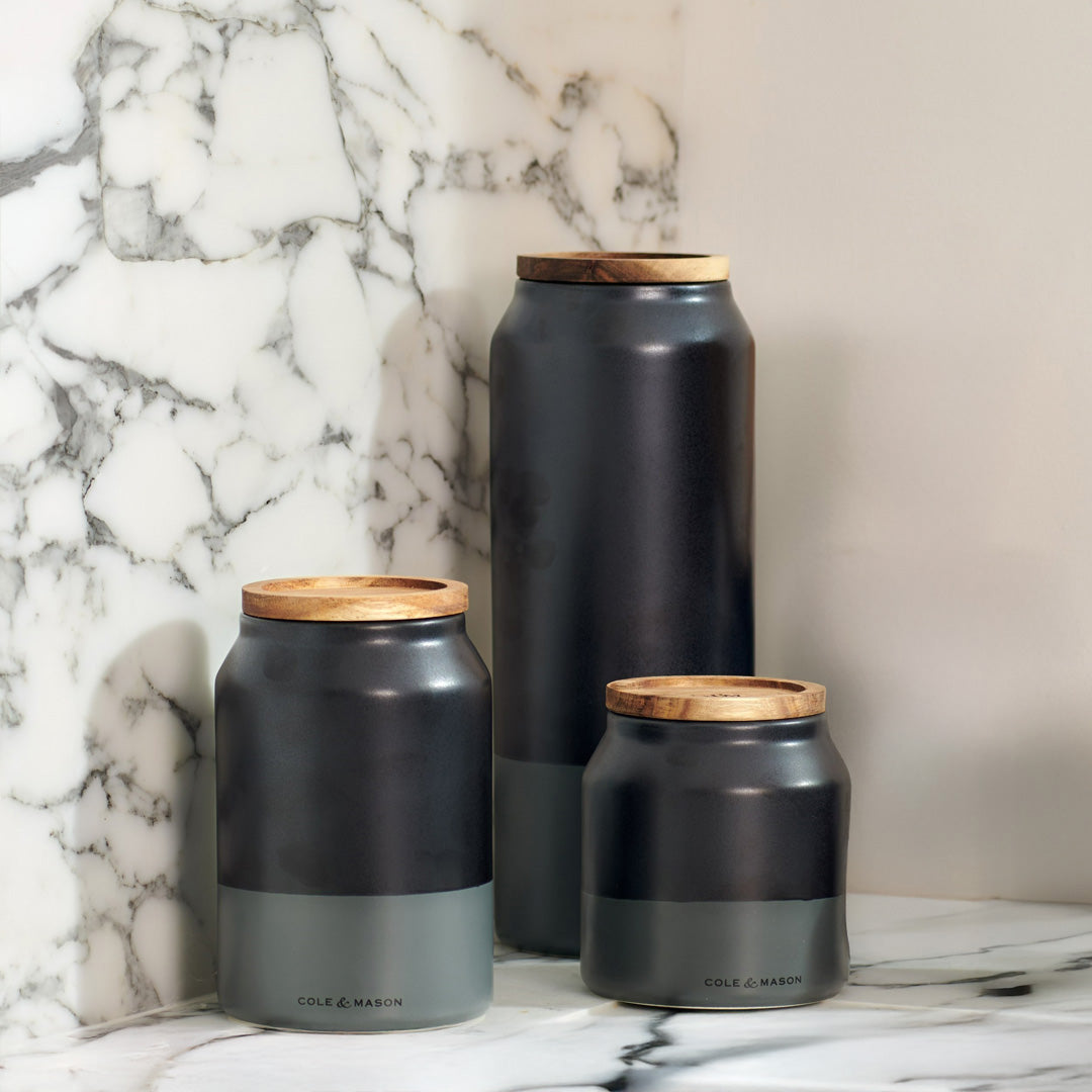Three matte black and gray ceramic canisters with wooden lids sit on a marble countertop against a marble wall. The canisters are labeled Cole & Mason and vary in height from small to large.