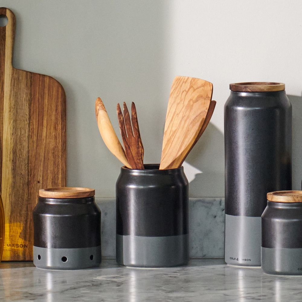 The Cole & Mason Linton Ceramic Utensil Pot, a matte black and gray soda can-shaped container with branding near the base, is ideal for storing cooking utensils and shown against a light beige background.