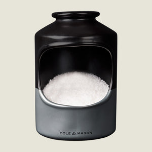 A ceramic salt cellar with a wide opening, filled with coarse sea salt. The container is black and gray with Cole & Mason printed on the front.