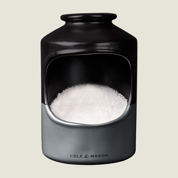 A ceramic salt cellar with a wide opening, filled with coarse sea salt. The container is black and gray with Cole & Mason printed on the front.