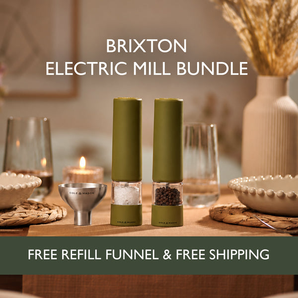 Two olive green Cole & Mason Brixton Electric Mills with a durable ceramic grinding system are displayed on a set dining table alongside plates, glasses, and a metal refill funnel. Text reads: Brixton Electric Mill Olive Green Set—Free Refill Funnel & Free Shipping. Convenient USB-C powered design.