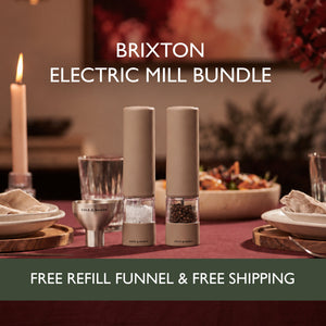 A dinner table featuring the Cole & Mason Brixton Electric Mill Stone Set—two rechargeable mills (one for salt, one for pepper) each with a robust ceramic grinding system—alongside a refill funnel, candles, and dishes. Text reads: BRIXTON ELECTRIC MILL STONE SET. FREE REFILL FUNNEL & FREE SHIPPING by COLE & MASON.
