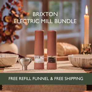 Two electric mills for salt and pepper—the Cole & Mason Brixton Electric Mill Terracotta Set—are displayed on a dining table set with plates, bread, and a candle, accompanied by a refill funnel. Text spotlights this bundle featuring USB-C rechargeable salt and pepper grinders with ceramic grinding systems from Cole & Mason, including free shipping and a refill funnel.
