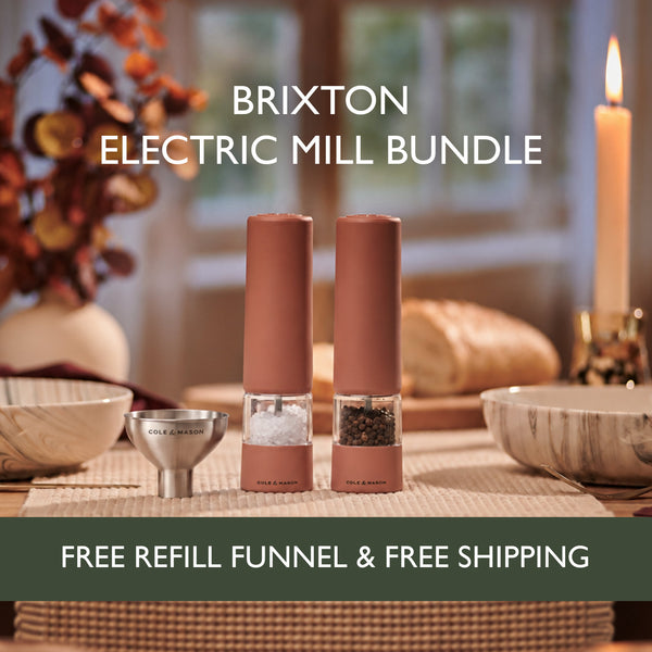 Two electric mills for salt and pepper—the Cole & Mason Brixton Electric Mill Terracotta Set—are displayed on a dining table set with plates, bread, and a candle, accompanied by a refill funnel. Text spotlights this bundle featuring USB-C rechargeable salt and pepper grinders with ceramic grinding systems from Cole & Mason, including free shipping and a refill funnel.