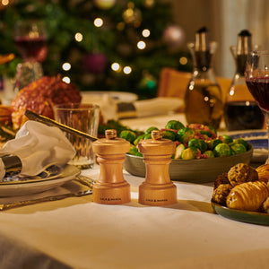 A festive dinner table with Brussels sprouts, roasted meat, bread rolls, wine, and two Cole & Mason Hoxton Natural Beech Mills, with a decorated Christmas tree in the background—perfect for a Christmas Gift Hamper.