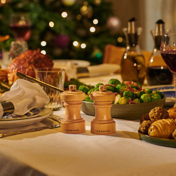 A festive dinner table with Brussels sprouts, roasted meat, bread rolls, wine, and two Cole & Mason Hoxton Natural Beech Mills, with a decorated Christmas tree in the background—perfect for a Christmas Gift Hamper.
