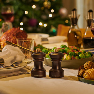 A festive table with Brussels sprouts and bread features a Cole & Mason Hoxton Chocolate Wood Mill, perfect for the Hoxton Chocolate Wood Mill Christmas Gift Hamper. A twinkling Christmas tree glows in the background.