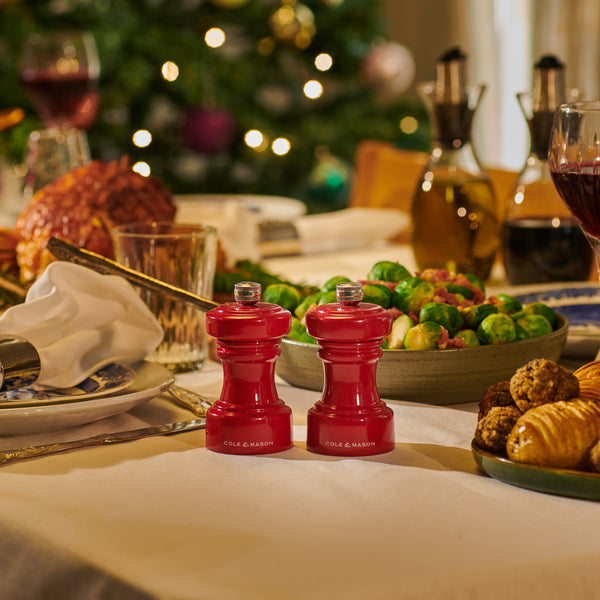 A festive table set with food, including Brussels sprouts and pastries, features Cole & Mason Hoxton Red Gloss Mills at the center; a blurred Christmas tree behind—perfect inspiration for the Hoxton Red Gloss Mill Christmas Gift Hamper.